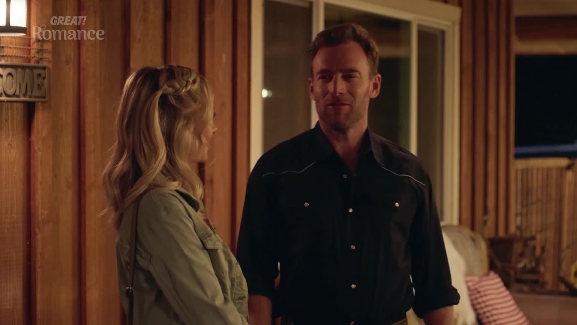 A man in a black shirt talks to a woman with braided blonde hair. They stand on a wooden porch, the air feeling warm and inviting.
