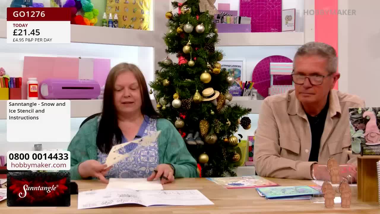 A woman holds up a stencil, demonstrating how to use it on a sheet of paper. Beside her, a man in glasses watches the demonstration on this Hobby Maker show, in the United Kingdom.
