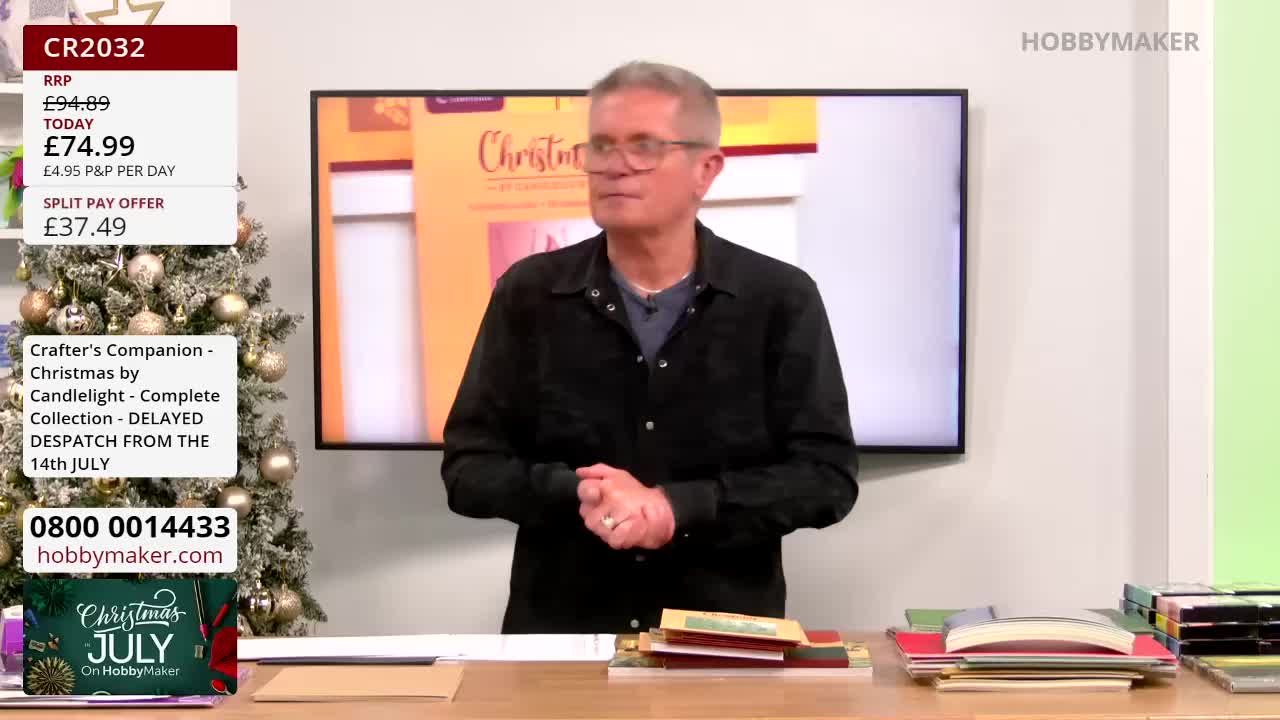 A man in glasses stands at a table, gesturing towards a display screen behind him.  He's on a Hobby Maker broadcast, and a Christmas-themed craft collection is being promoted.
