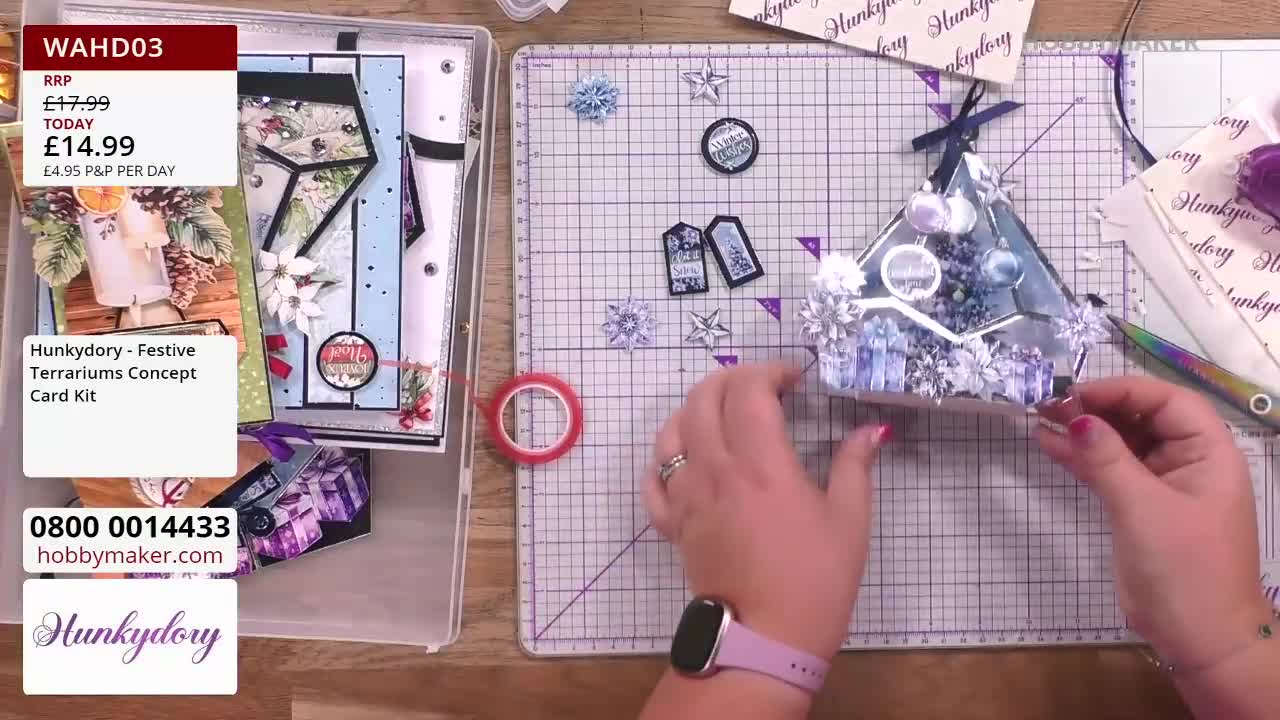 A woman carefully positions a small, decorative terrarium, part of a card kit from Hobby Maker in the United Kingdom.  Tiny paper snowflakes and tags are scattered on the cutting mat, ready to be added to the miniature scene.
