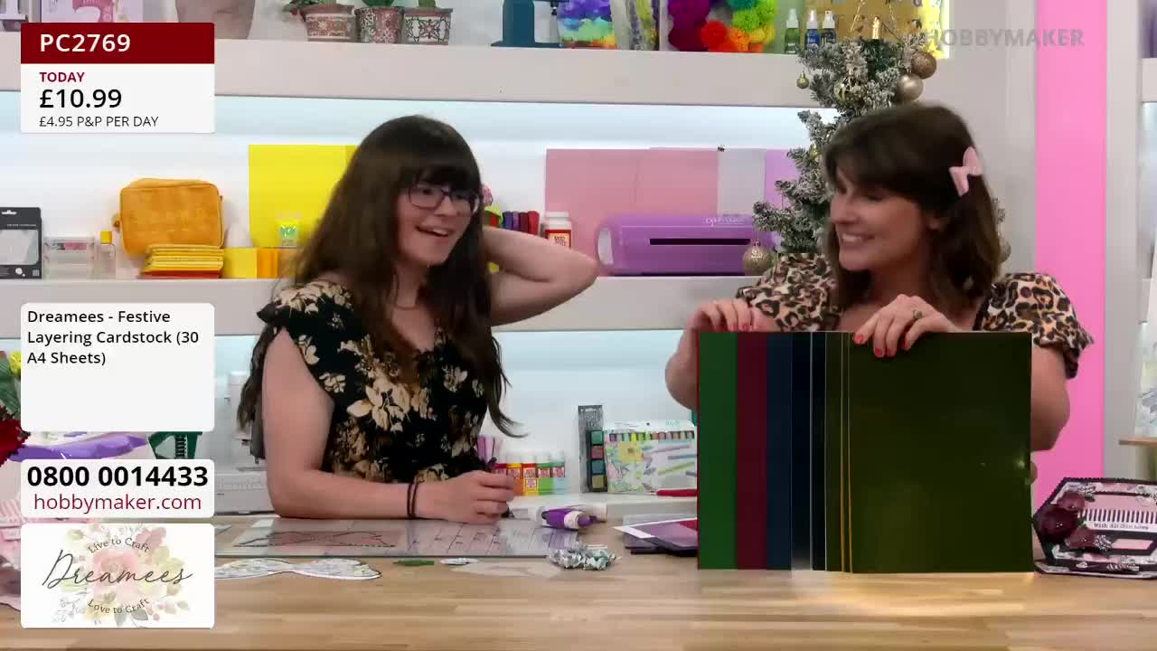 Two women at a Hobby Maker demonstration are showing off a stack of colored cardstock. One woman, wearing a leopard-print top, holds the cardstock up while the other, smiling, gestures towards it.
