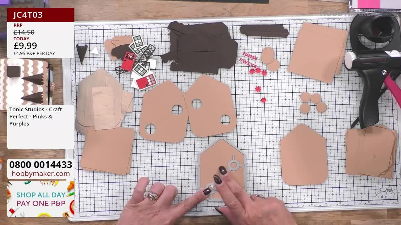 A crafter, likely from the United Kingdom, carefully presses a small die-cut shape onto a beige paper house. Various pre-cut elements, including windows and roof pieces, are scattered around the workspace.
