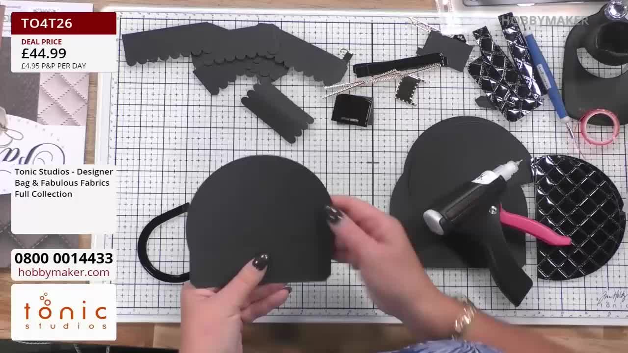 A woman holds a curved black piece of cardstock, seemingly about to glue it to another piece shaped like a half-circle. Scattered around the work surface are various black cutouts and a Hobby Maker hot glue gun.
