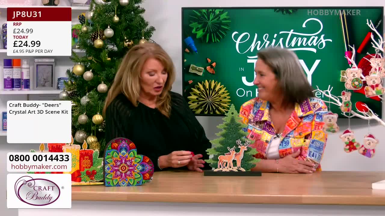 Two women are seated at a table, discussing a "Craft Buddy" crystal art kit. The scene is set in a studio for the UK's Hobby Maker, with Christmas decorations in the background.

