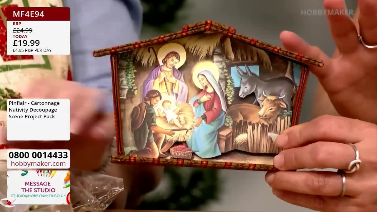 A person's hands hold a small, layered nativity scene, likely a Hobby Maker craft project. The colorful, detailed artwork depicts the holy family and animals in a stable.
