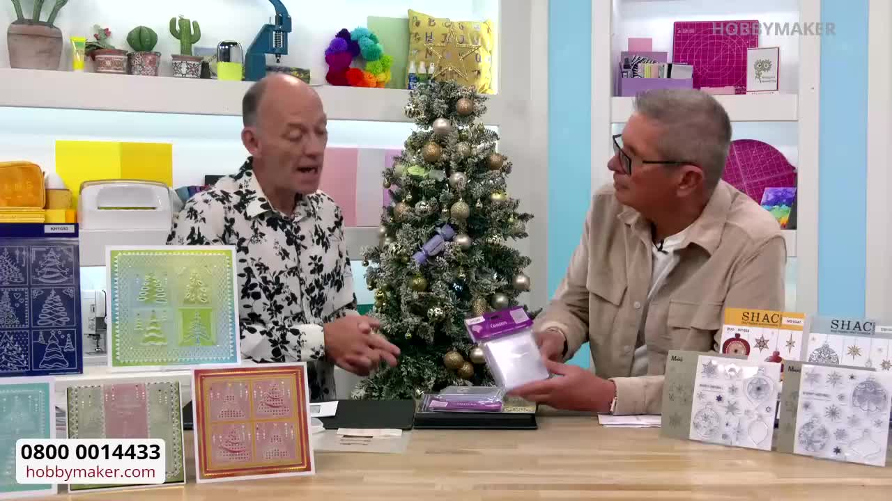 Two men are seated at a table, discussing crafting supplies in a brightly lit studio, likely for a segment on Hobby Maker. One gestures towards samples of embossed cards, while the other holds up a package of clear stamps.
