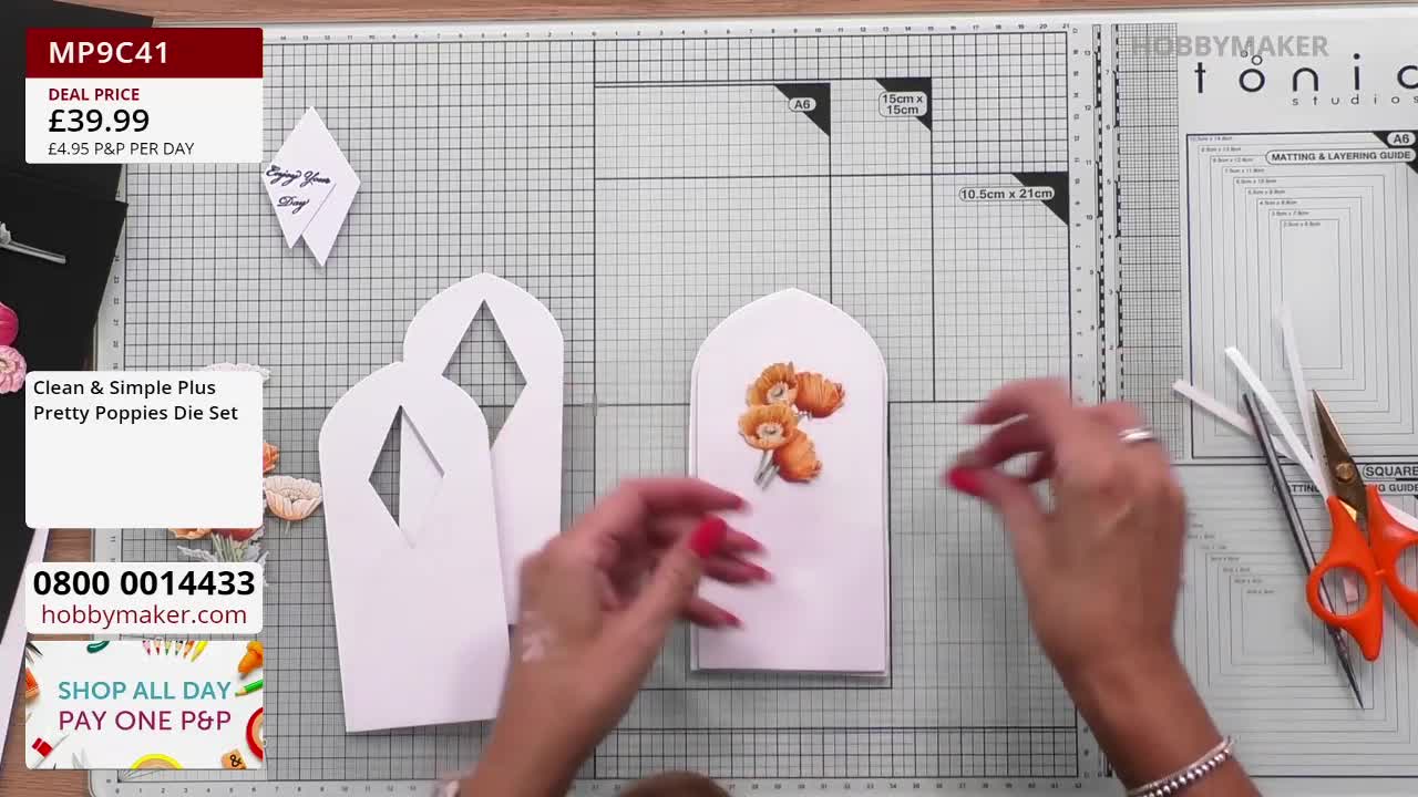 On a Hobby Maker studio surface, a woman's hands are arranging paper cutouts of white arched shapes. She's placing small, printed poppy images onto the central card.
