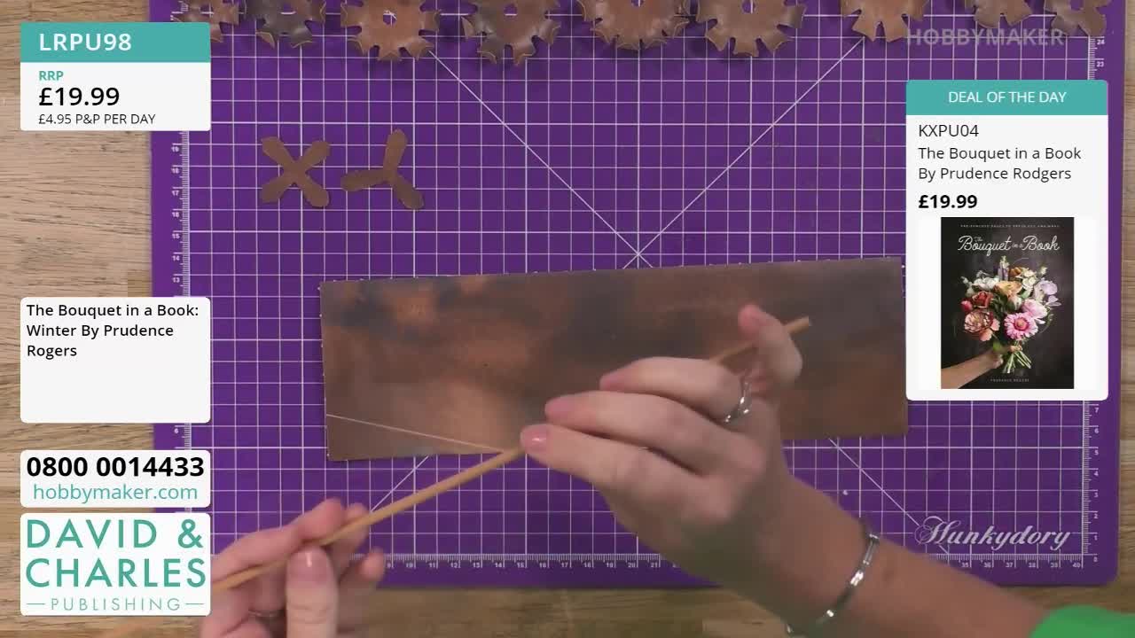 A woman's hands hold a thin wooden dowel, working with a piece of copper-colored material on a purple cutting mat. The Hobby Maker logo is visible in the background, along with a book cover featuring a floral bouquet.
