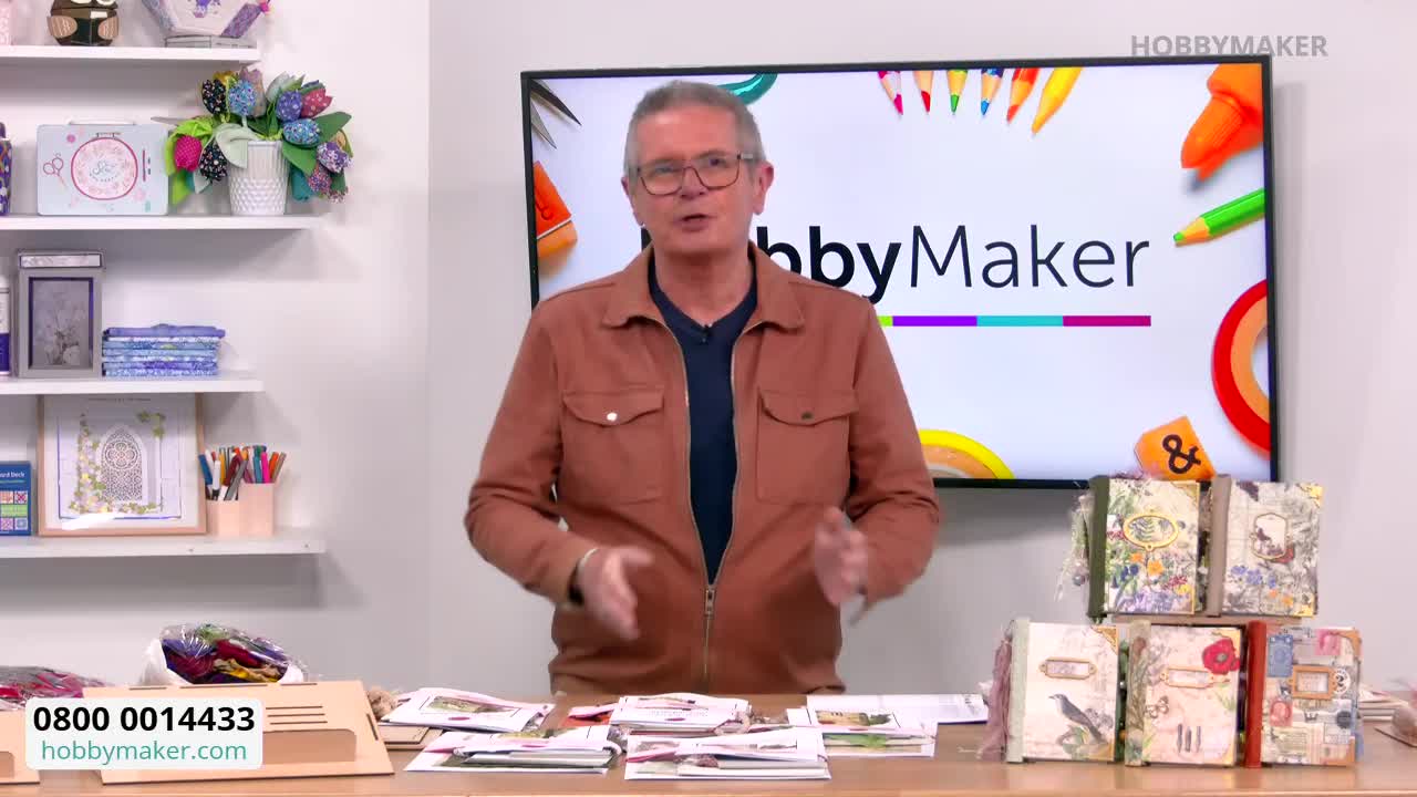 A man in a brown jacket gestures with his hands in front of a HobbyMaker backdrop. Several decorated books are stacked on the table in front of him.
