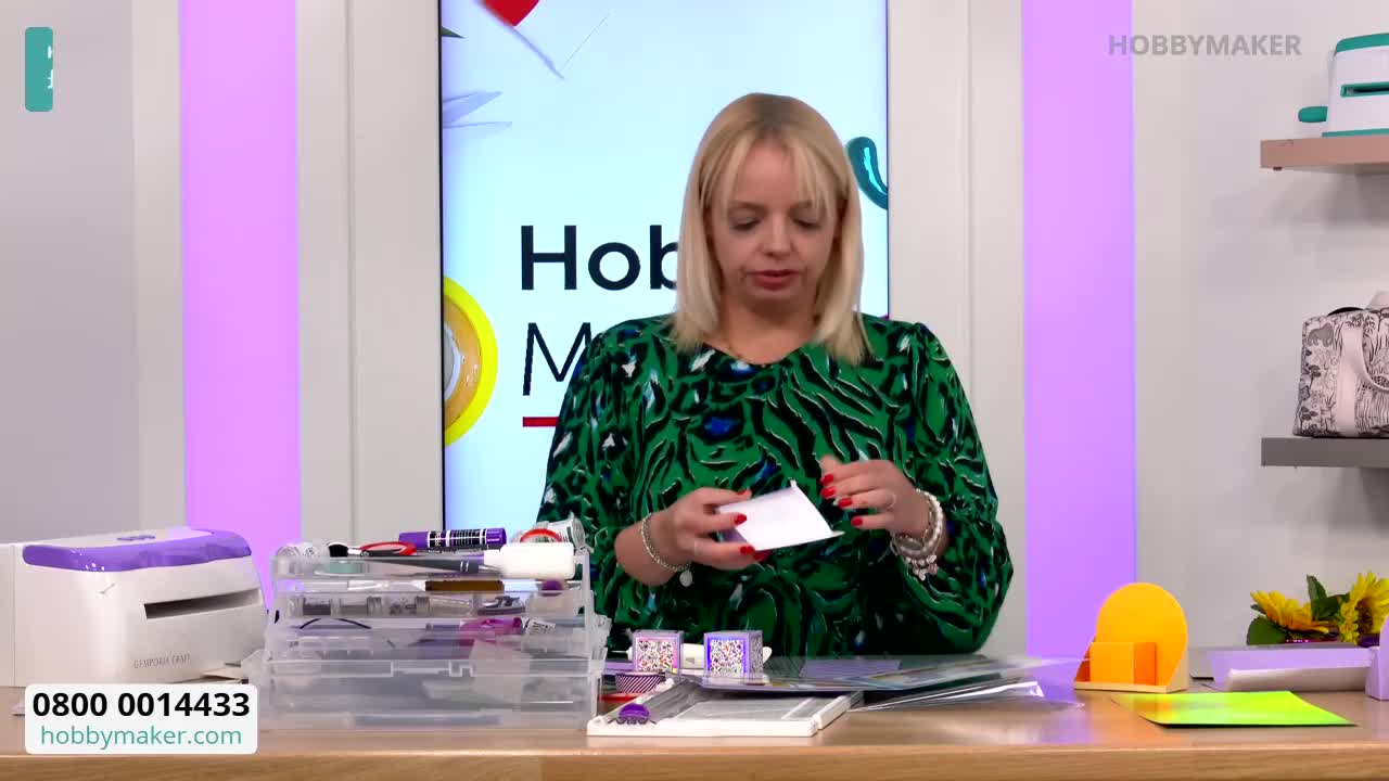The woman in the green patterned top is holding a small white card, carefully folding it. Behind her, the Hobby Maker logo is visible on a large screen.
