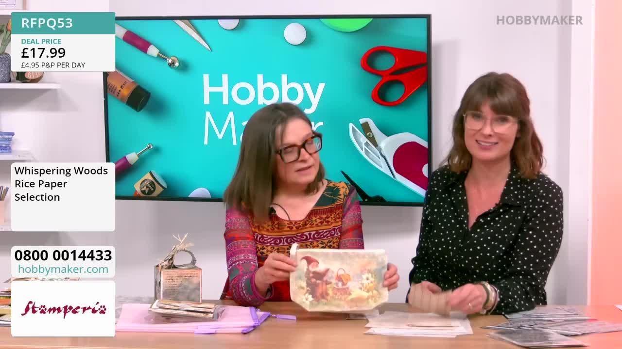 Two women are seated at a table, one holding up a decorated pouch while the other smiles at the camera. The backdrop features the Hobby Maker logo and craft supplies, suggesting a demonstration from the United Kingdom.
