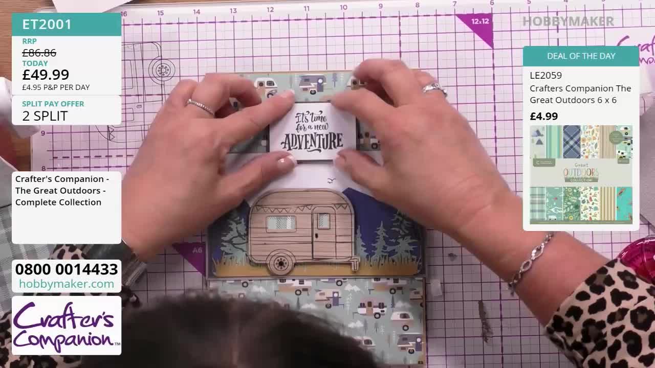 A woman's hands carefully position a small, rectangular sign that reads "It's time for a new adventure" above a paper camper van. The Hobby Maker logo and product details are visible in the background.
