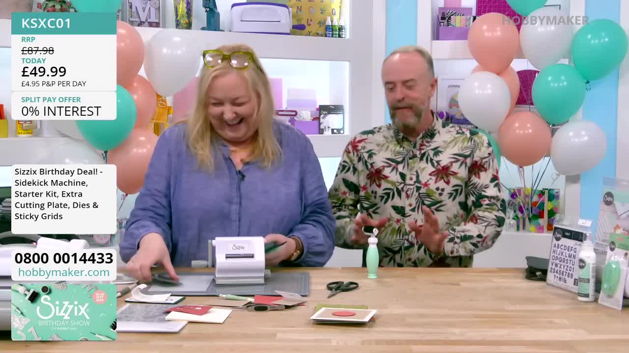 A woman with glasses is smiling, holding a small crafting machine. The man next to her gestures with his hands, and a cluster of balloons floats behind them.
