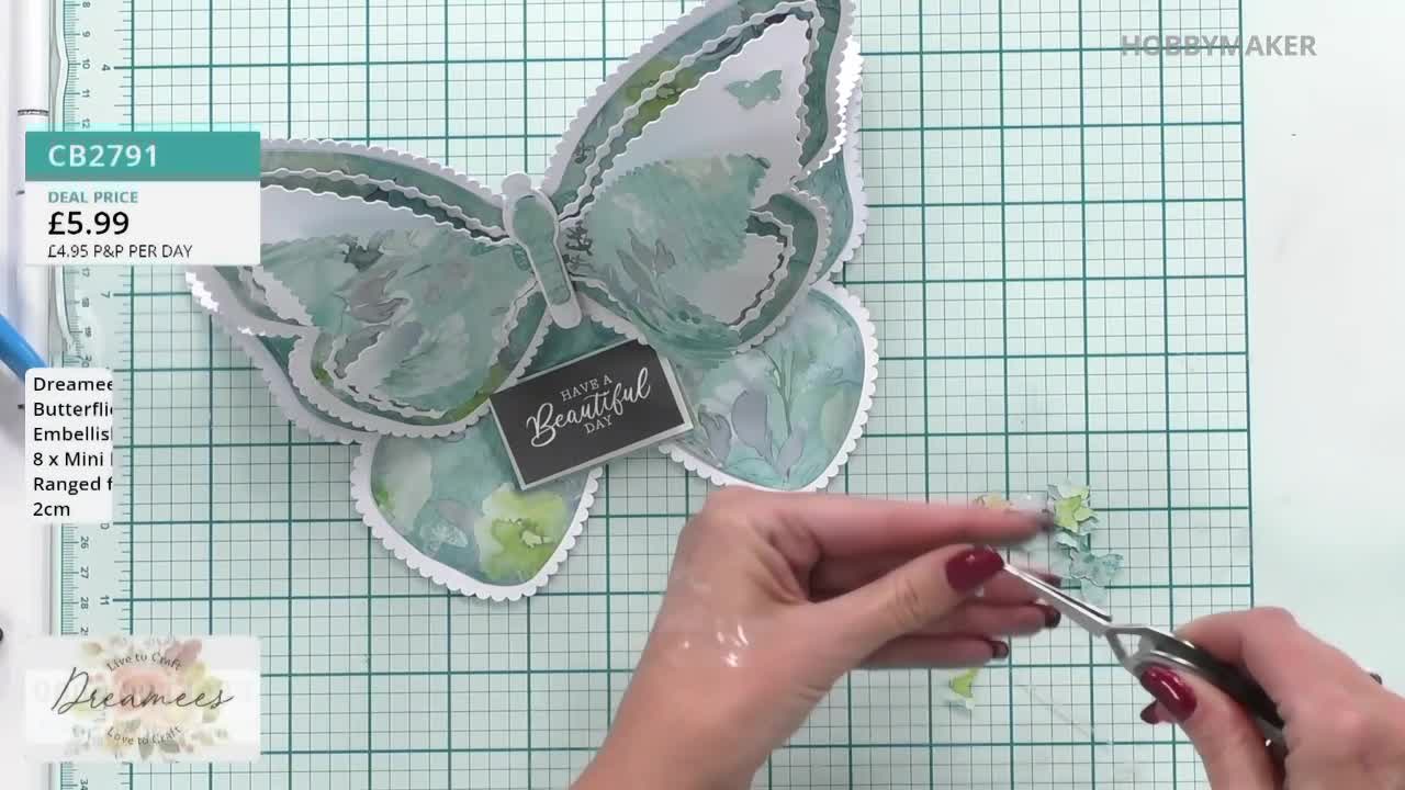 A woman is meticulously snipping tiny paper embellishments with scissors, preparing them for a butterfly-shaped card. The Hobby Maker product, a Dreamer butterfly, is nearly complete on the gridded surface.
