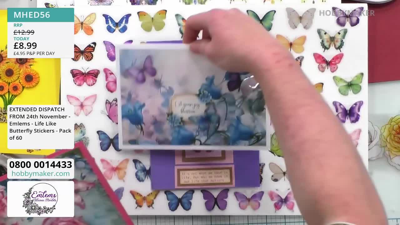 A hand lifts a card decorated with blue flowers and butterflies, revealing a purple base with a message. The Hobby Maker product display features butterfly stickers and a price tag.
