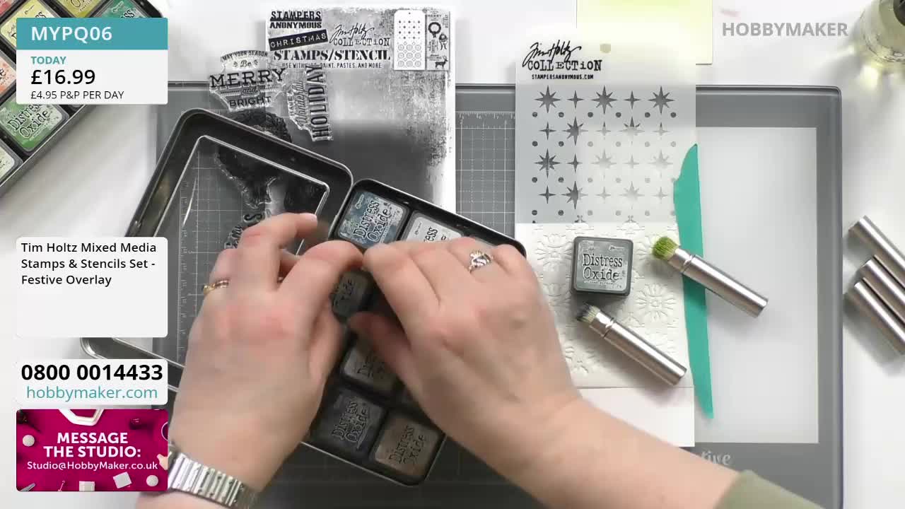 A person on Hobby Maker is opening a metal tin of ink pads, selecting one with their fingers. Beside the tin, a stencil and a small ink applicator are ready for use.
