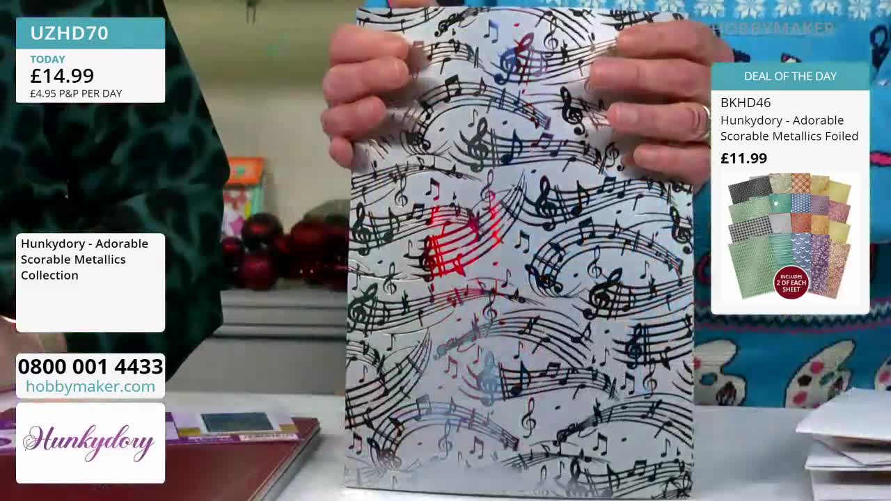 A person holds up a sheet of paper from Hobby Maker, covered in musical notes. The paper has a shiny, metallic finish, with some areas reflecting a red hue.
