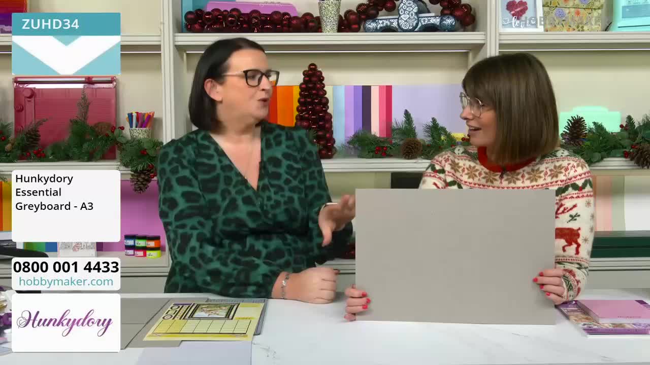 Two women are holding a large sheet of greyboard, discussing it on a Hobby Maker broadcast from the United Kingdom. One gestures with her hand as she speaks, while the other holds the board up for the camera.
