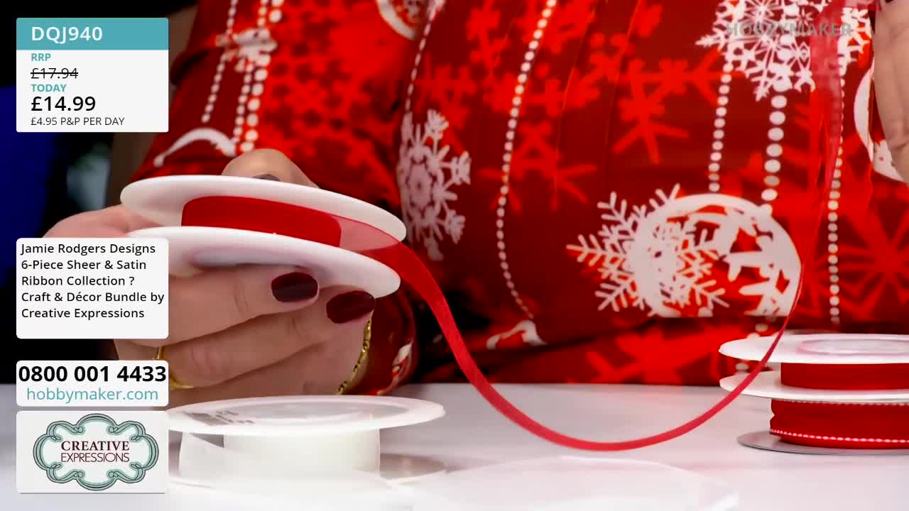 A hand holds a spool of red ribbon, pulling a length of it into the air. The ribbon is being demonstrated as part of a Hobby Maker craft bundle from the United Kingdom.

