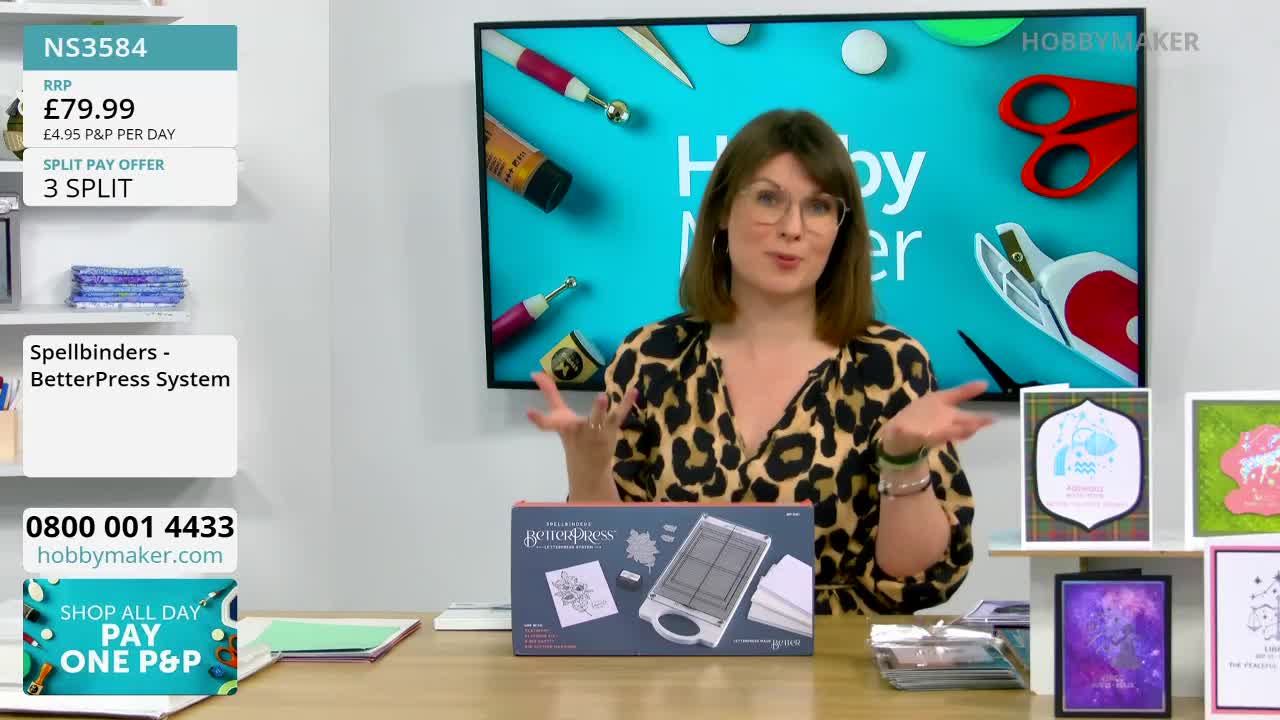 A woman in a leopard-print top gestures enthusiastically as she presents a crafting tool. On the table before her, a box for the Spellbinders BetterPress System sits next to several finished cards.