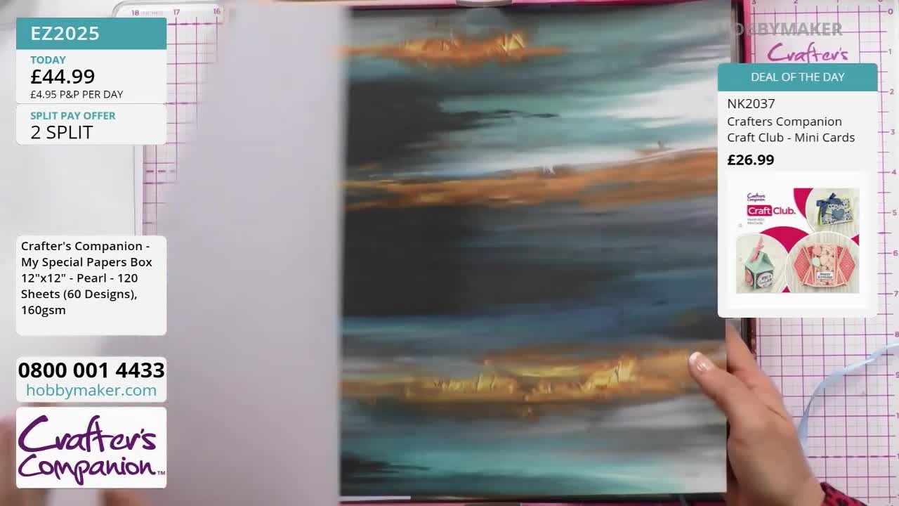 A hand turns a large sheet of paper with a dark, abstract design featuring streaks of teal and gold. Another hand holds a smaller stack of papers, showcasing a selection of card-making kits.