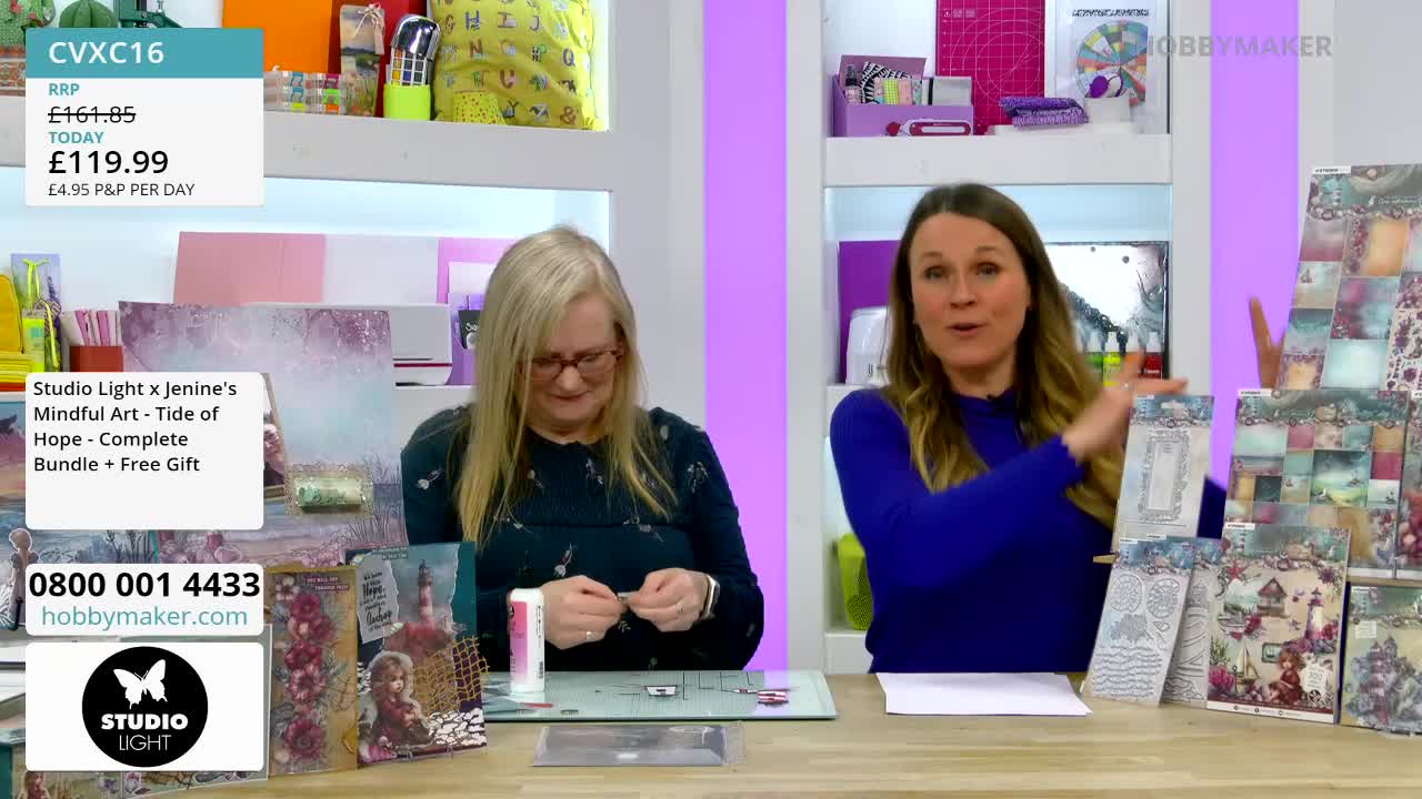A woman in a blue top gestures towards stacks of patterned paper and card-making supplies. Beside her, another woman carefully works on a craft project at a table.