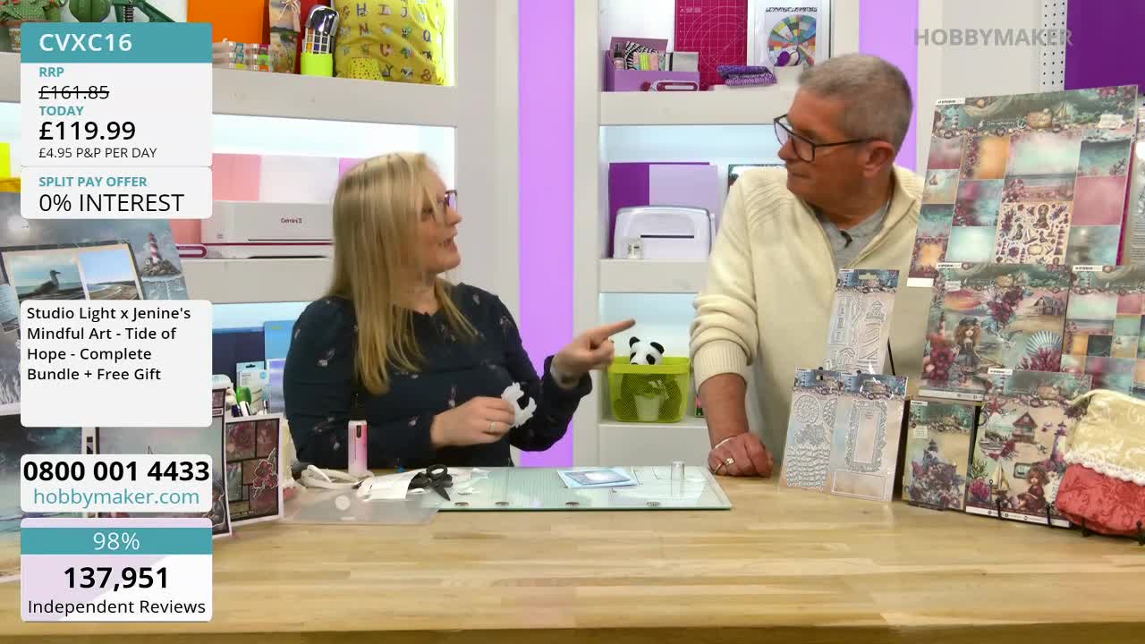 A woman with blonde hair gestures animatedly towards a table displaying crafting supplies. Beside her, a man in a cream jumper looks on, with stacks of patterned paper and stencils arranged in front of him.