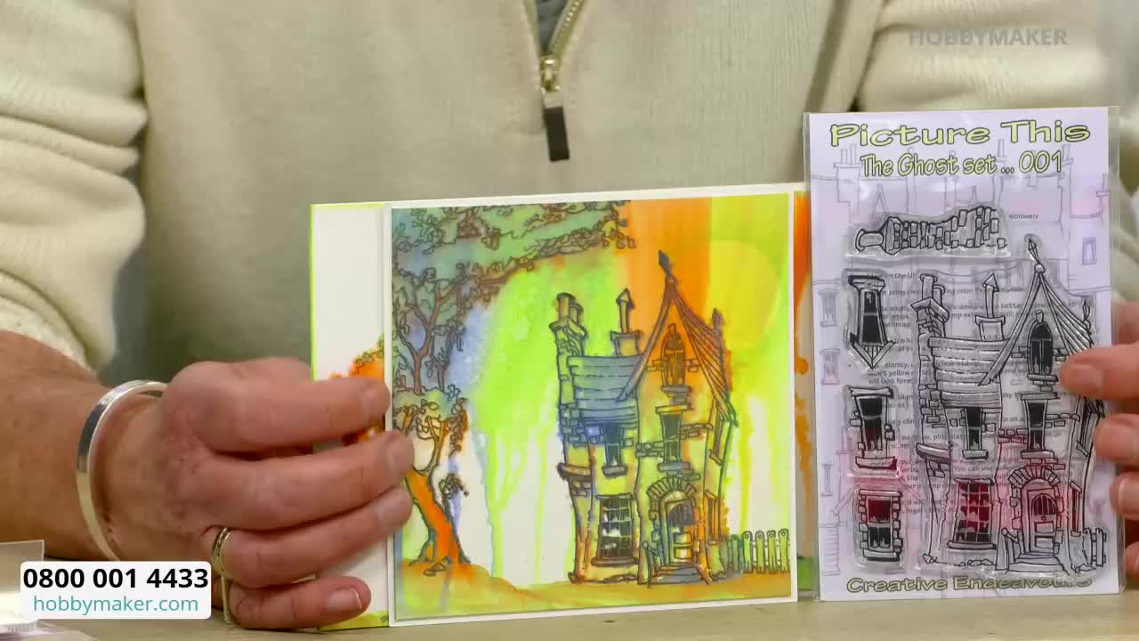 A hand holds up a card featuring a watercolour painting of a spooky house. Next to it, another hand displays a clear stamp set with the same house design.