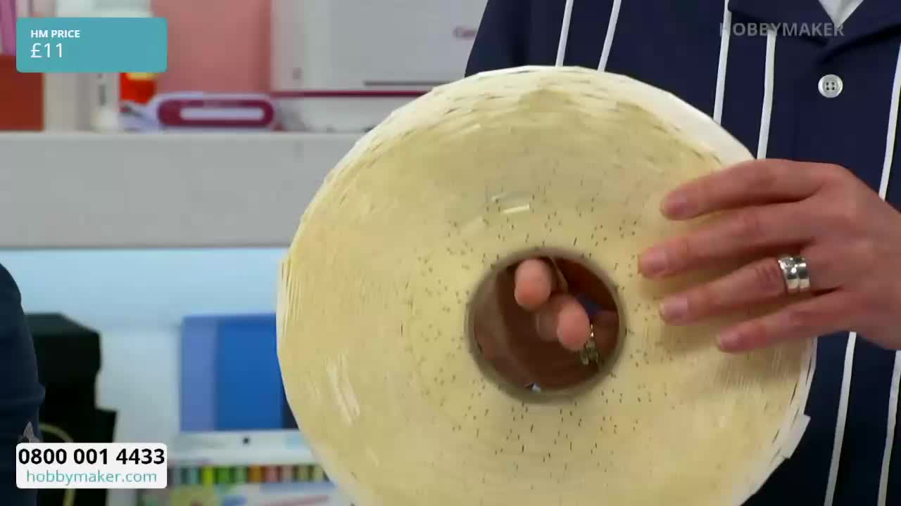 A person holds a large roll of pale yellow paper, its surface dotted with tiny black flecks. The HobbyMaker logo is visible on their striped shirt.