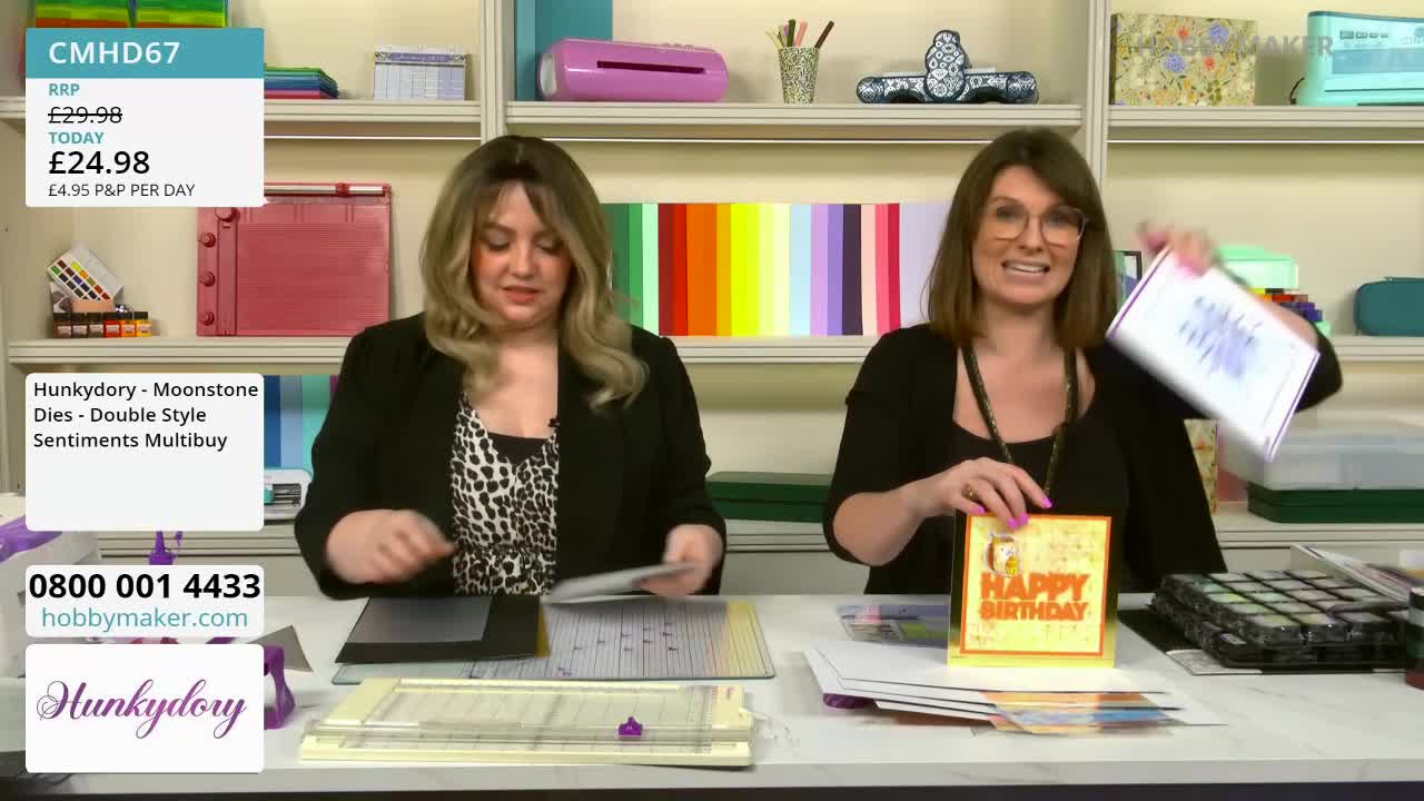 A woman holds up a finished card, its "Happy Birthday" message in bold orange letters. Beside her, another woman is placing a sheet of paper onto a cutting mat.