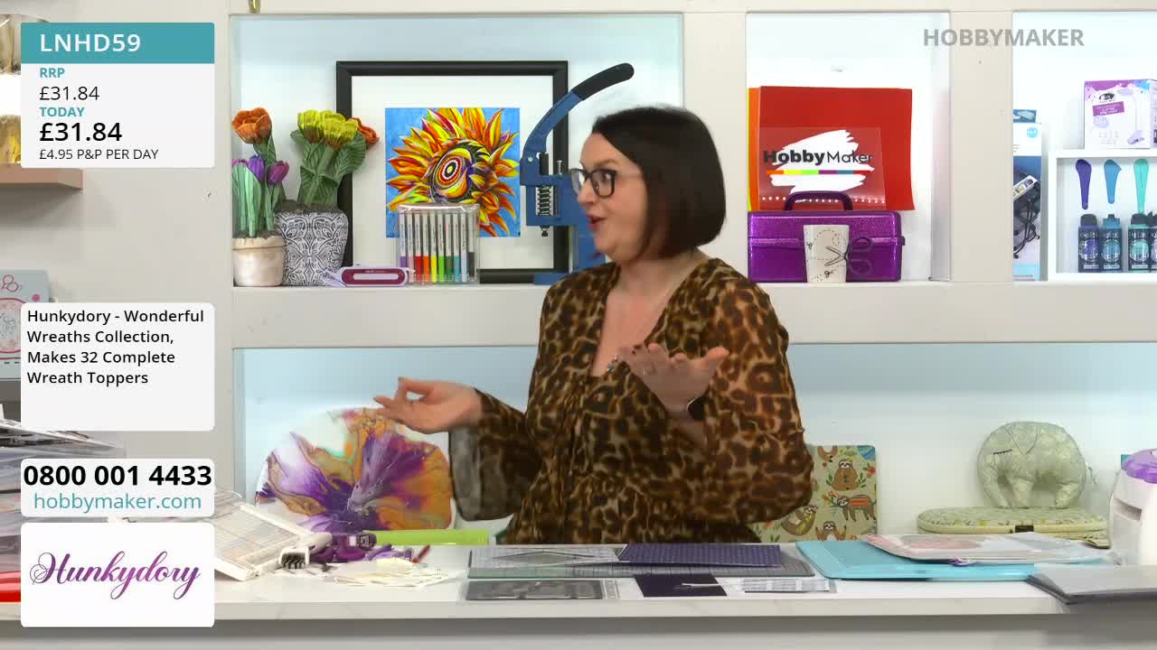 A woman in a leopard-print top gestures animatedly while discussing craft supplies. On the table before her, a colourful floral design is being worked on.