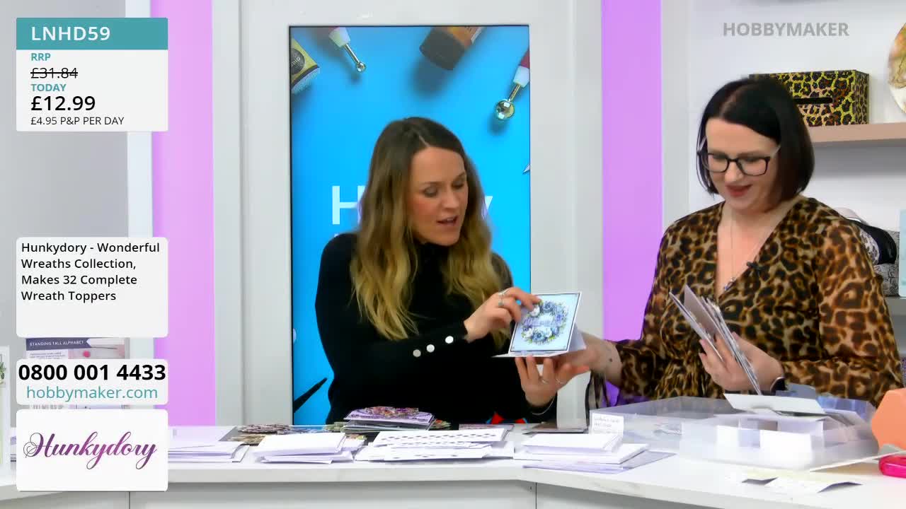 A woman in a black top holds up a small, decorated card, showing it to another woman in a leopard-print dress. The second woman smiles, looking at papers she holds in her hands.