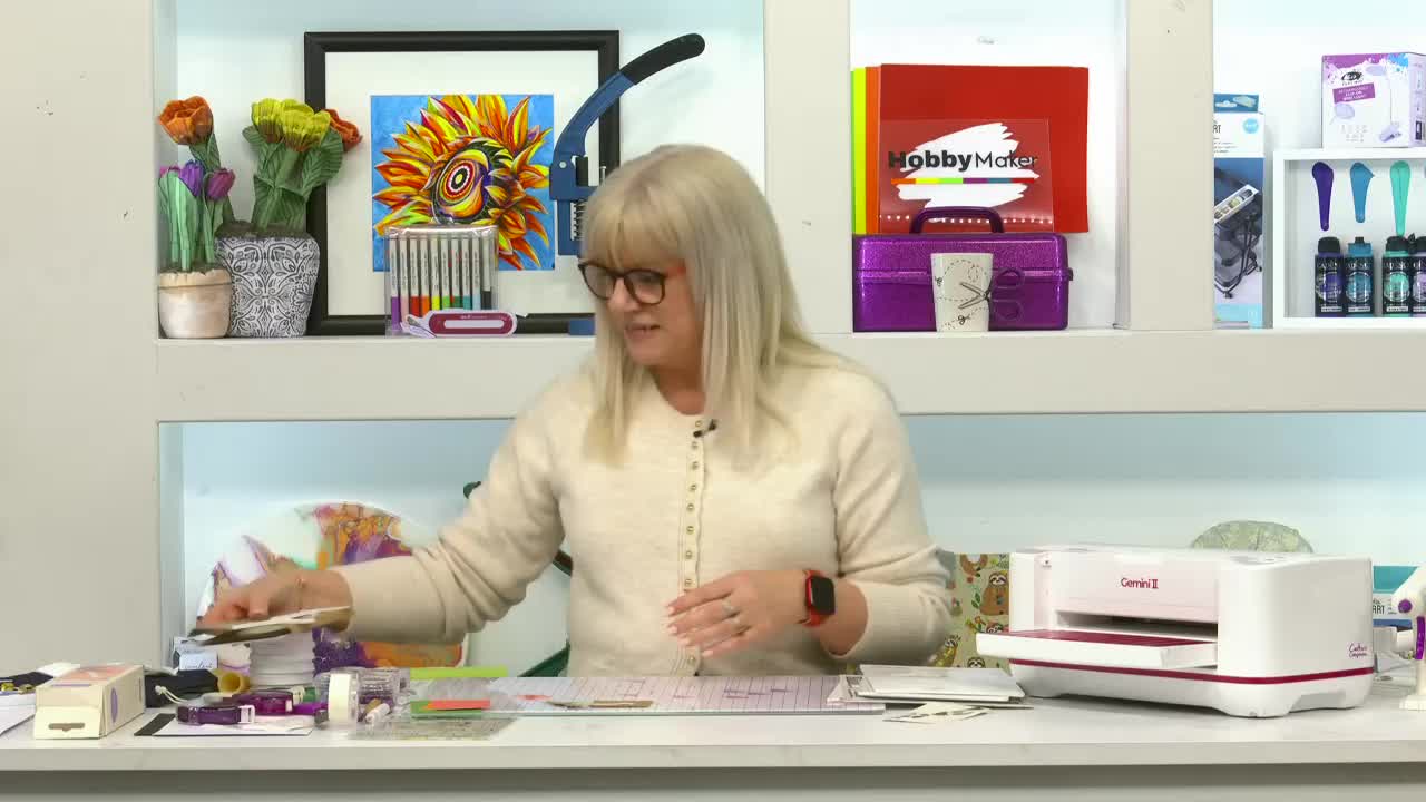 A woman in a cream cardigan is reaching for a stack of paper on a craft table. A white Gemini machine sits to her right, ready for use.