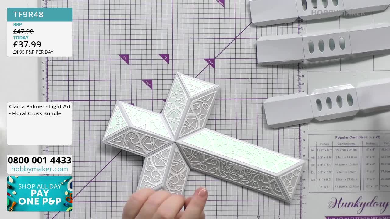 A delicate, white cross crafted from cardstock sits on a cutting mat.  Its intricate floral cutouts catch the light as a hand gently adjusts one of its panels.