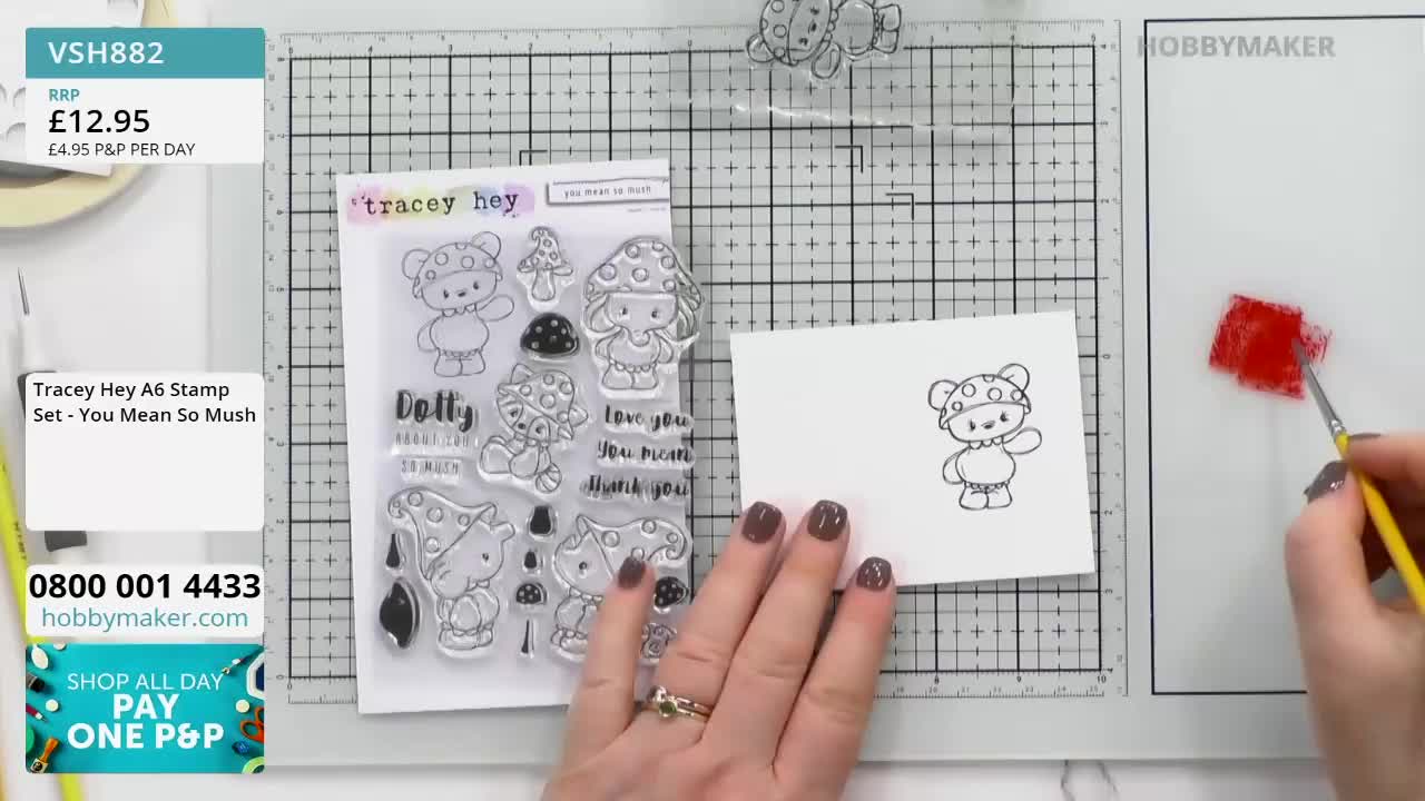 A hand with a yellow-handled brush is dabbing red paint onto a white surface. Nearby, a small card features a stamped image of a teddy bear wearing a mushroom hat.