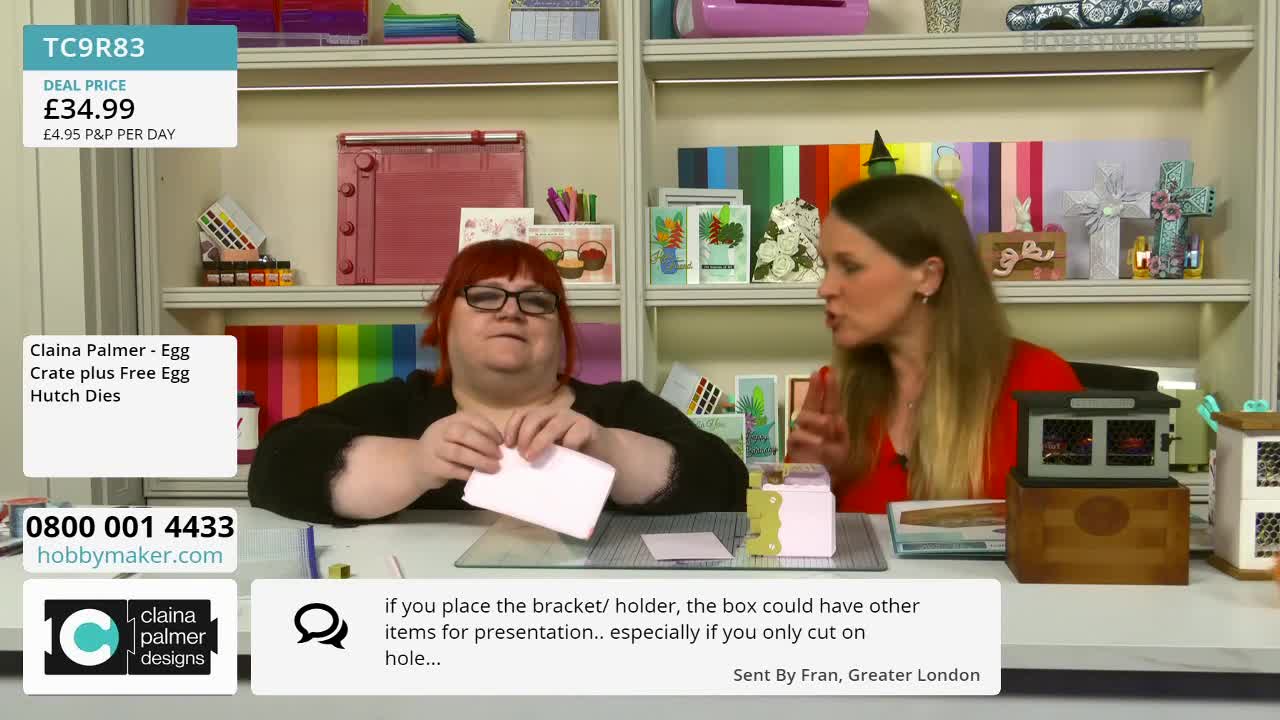 A woman with red hair and glasses is folding a small pink card. To her right, another woman in a red top gestures with her hands as she speaks.