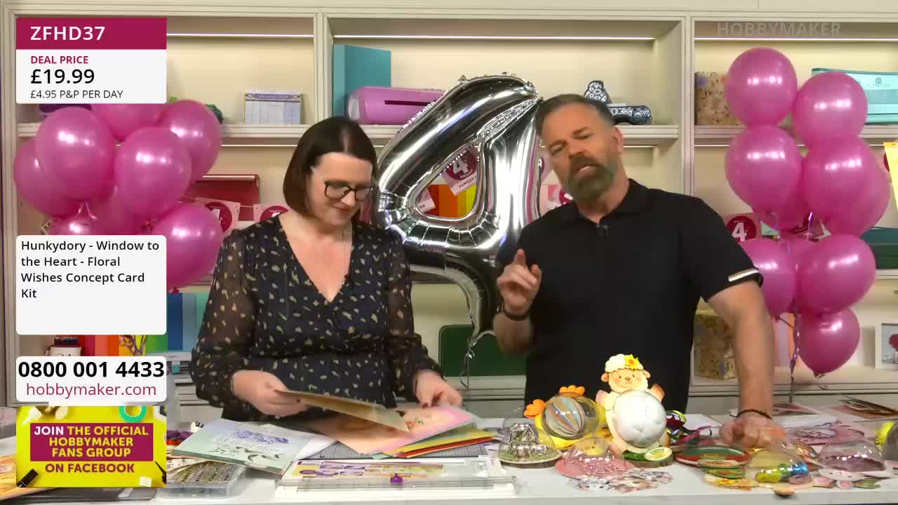 A man with a beard gestures enthusiastically while a woman sorts through card-making supplies. Pink balloons cluster behind them, and a large silver "4" balloon looms over the scene.