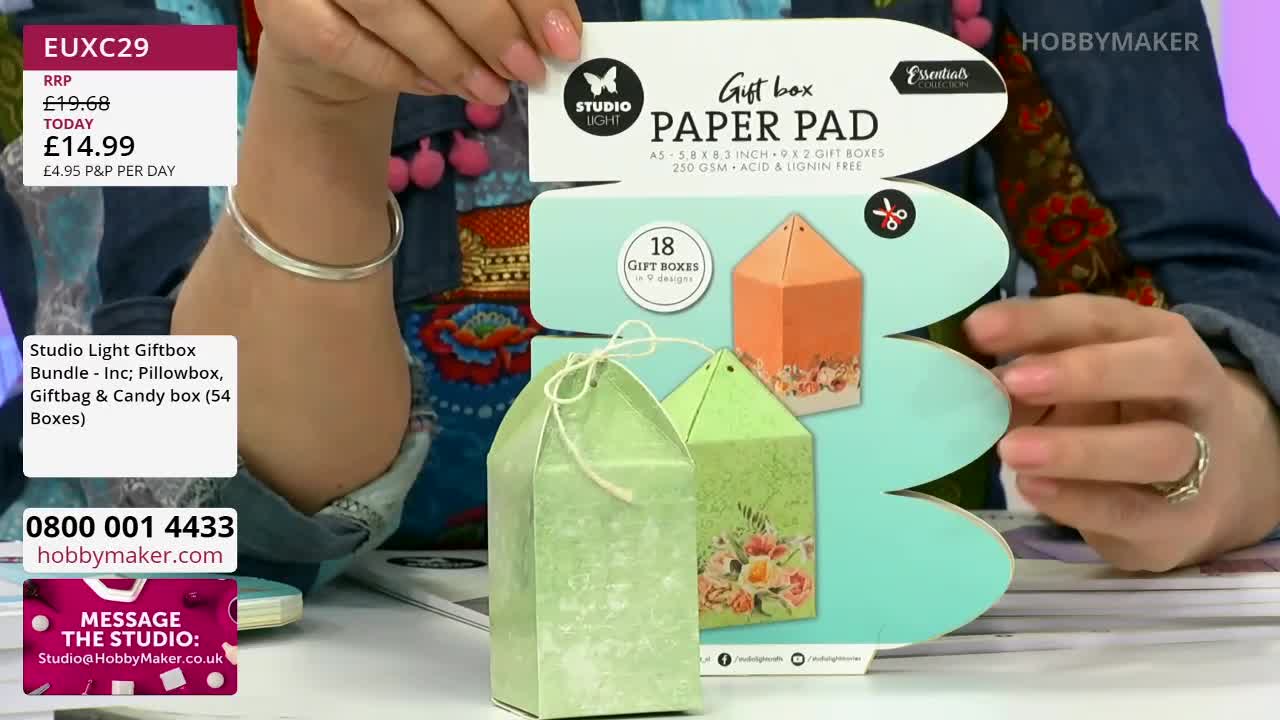 Two decorative gift boxes, one pale green and the other a soft orange, stand on a table. A woman's hands, adorned with a silver ring, hold up a large card featuring images of these boxes.