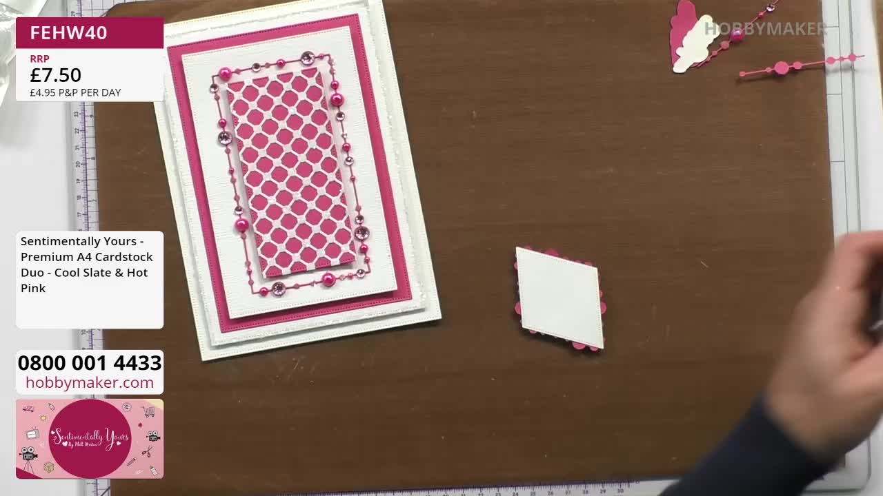 A hand reaches in to place a small, diamond-shaped piece of cardstock, adorned with tiny pink circles, onto a brown crafting mat. Beside it, a finished card, featuring a pink patterned insert and delicate pearl embellishments, sits ready.