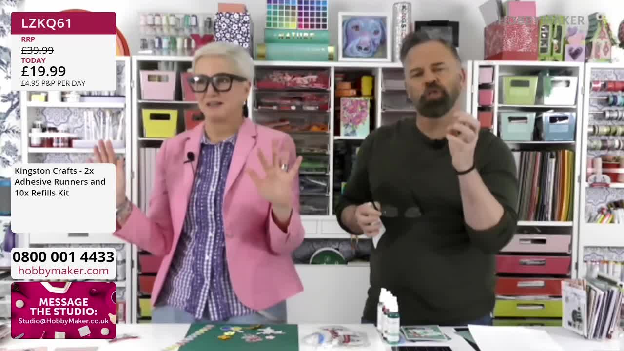 A woman in a pink blazer gestures with both hands while a man in a dark green shirt holds up a small item between his thumb and forefinger. They stand in front of shelves stocked with craft supplies. A woman in a pink blazer gestures with both hands while a man in a dark green shirt holds up a small item between his thumb and forefinger. They stand in front of shelves stocked with craft supplies.