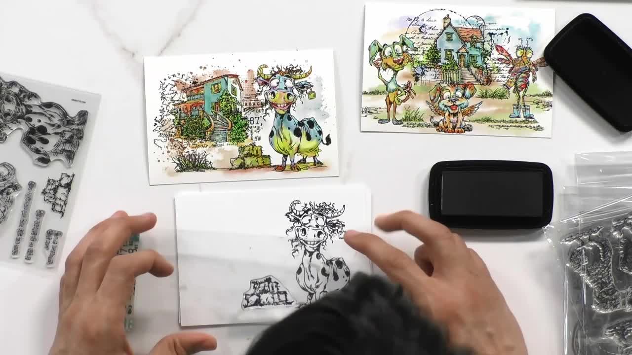 My hands hold a clear stamp, ready to press it onto the card. Two finished watercolour cards, one with a whimsical cow and another with woodland creatures, sit nearby. My hands hold a clear stamp, ready to press it onto the card. Two finished watercolour cards, one with a whimsical cow and another with woodland creatures, sit nearby.
