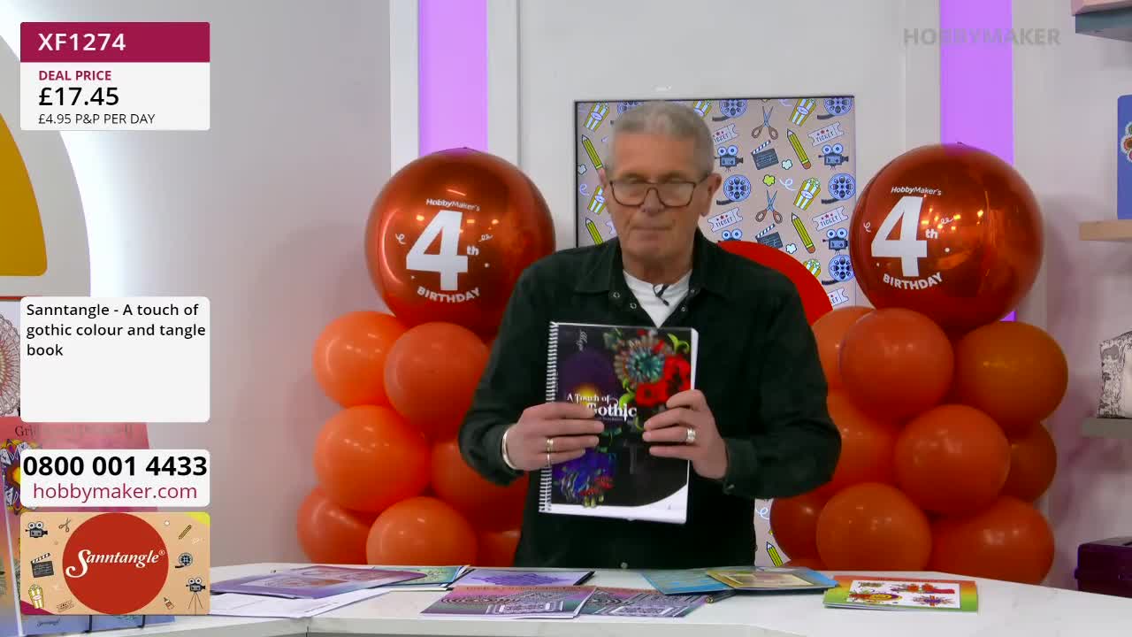 A man holds up a book titled "A touch of gothic colour and tangle," with a stack of orange balloons behind him. The Hobby Maker logo is visible on a large balloon. A man holds up a book titled "A touch of gothic colour and tangle," with a stack of orange balloons behind him. The Hobby Maker logo is visible on a large balloon.