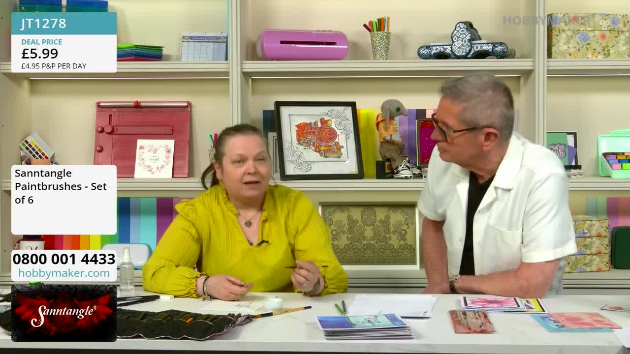 A woman in a bright yellow top gestures with a fine-tipped pen as she speaks to a man in a white shirt. On the table before them, a stack of colourful cards and art supplies are laid out.