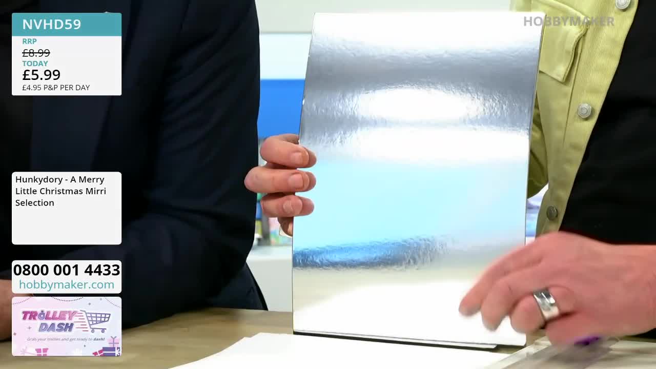 A hand holds up a sheet of shimmering silver cardstock. The card, part of a "Merry Little Christmas Mirri Selection" from Hobby Maker, reflects the bright studio lights.