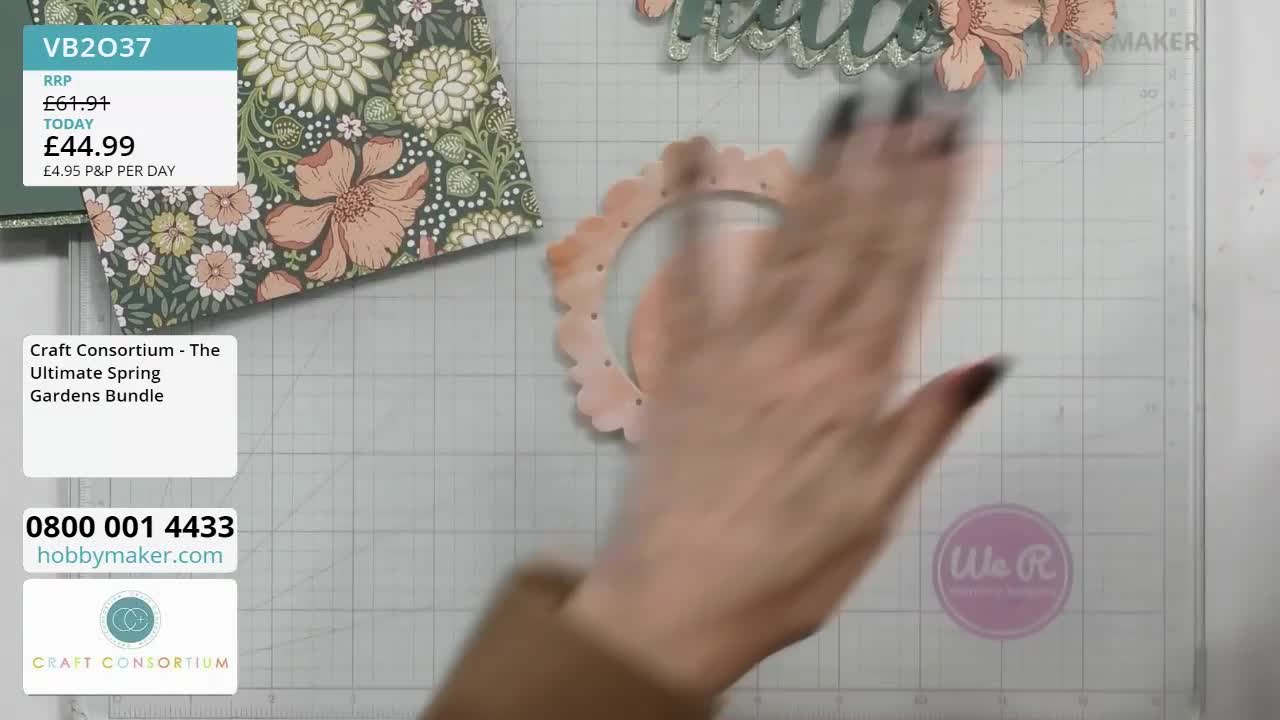 A hand sweeps across a delicate, scalloped pink frame, obscuring it from view. Nearby, a sheet of patterned paper featuring bold floral designs lies ready for crafting.