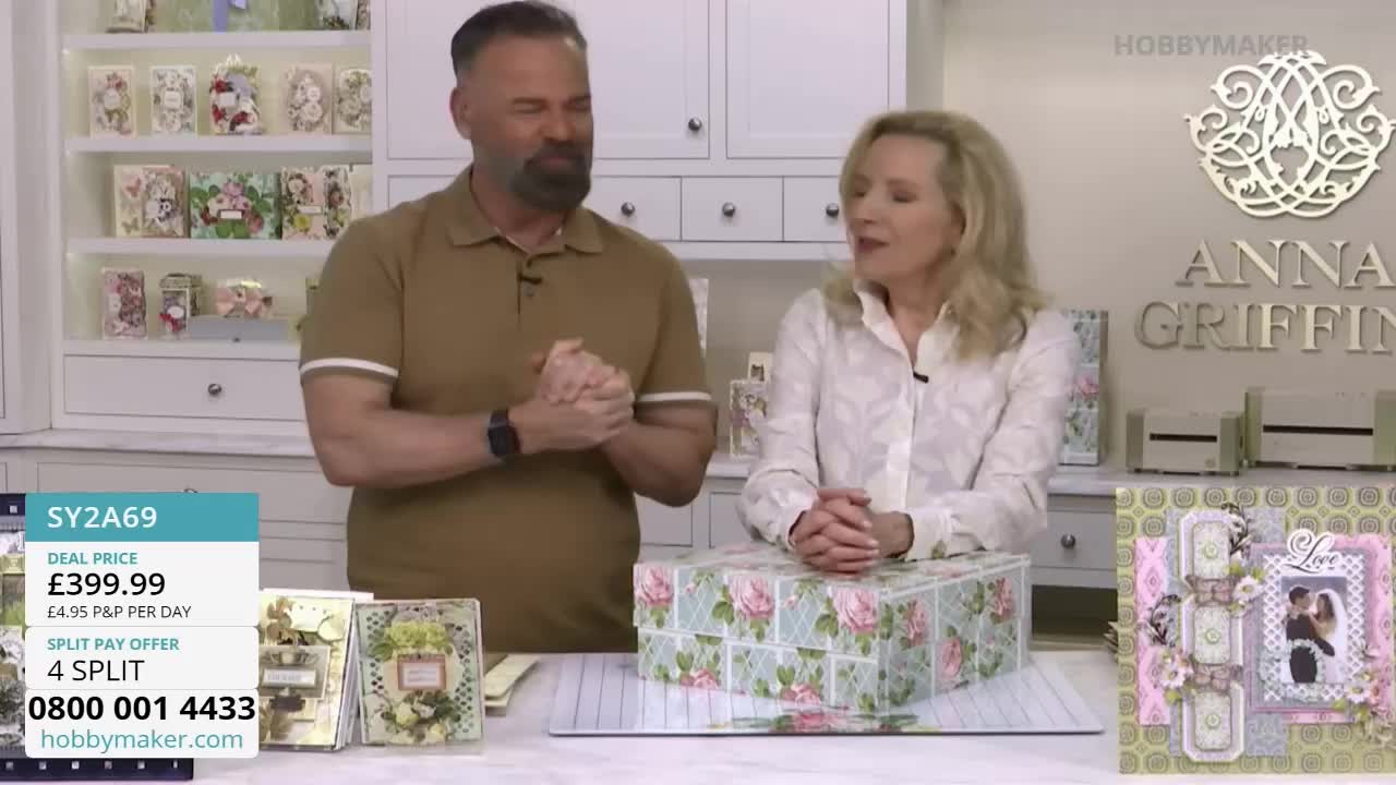 A man and a woman stand behind a table displaying a large, floral-patterned box. Shelves filled with crafting supplies are visible in the background.