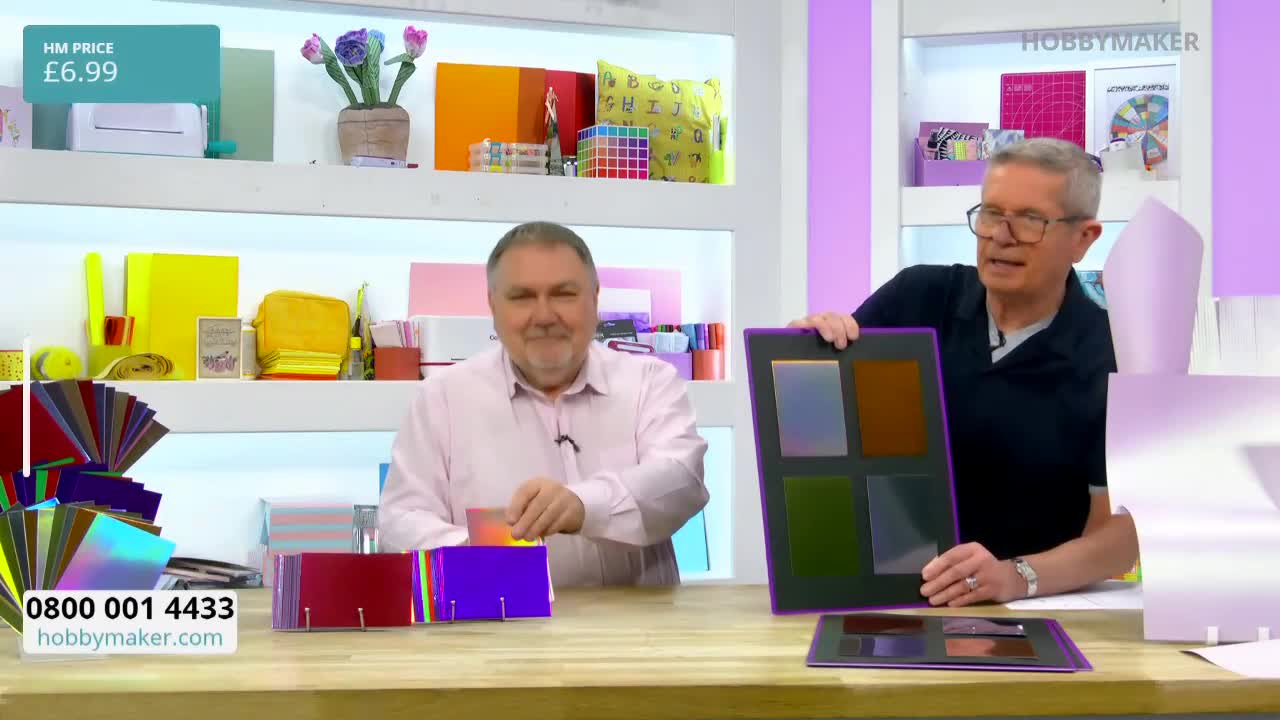 A man in a pink shirt carefully places a small, iridescent card into a purple holder. Beside him, another man holds up a larger display board featuring four swatches of coloured paper.
