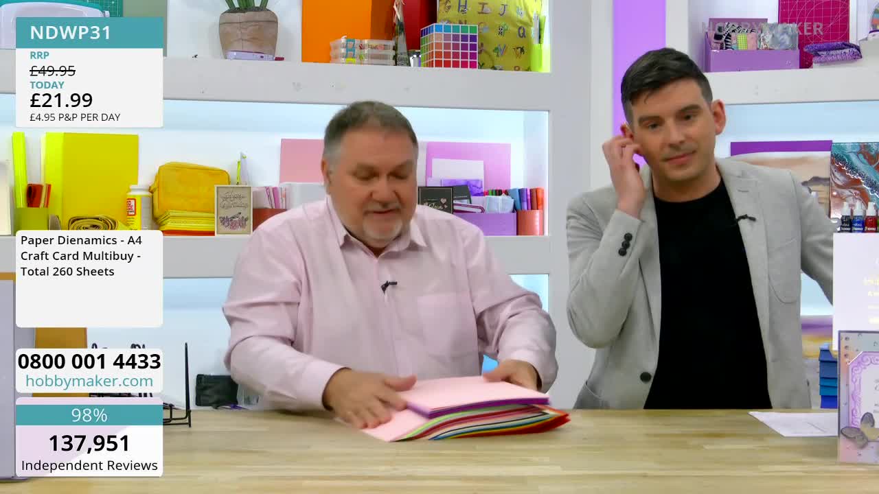 A man in a pink shirt is fanning out a stack of colourful cardstock. Beside him, another man is holding a phone to his ear.