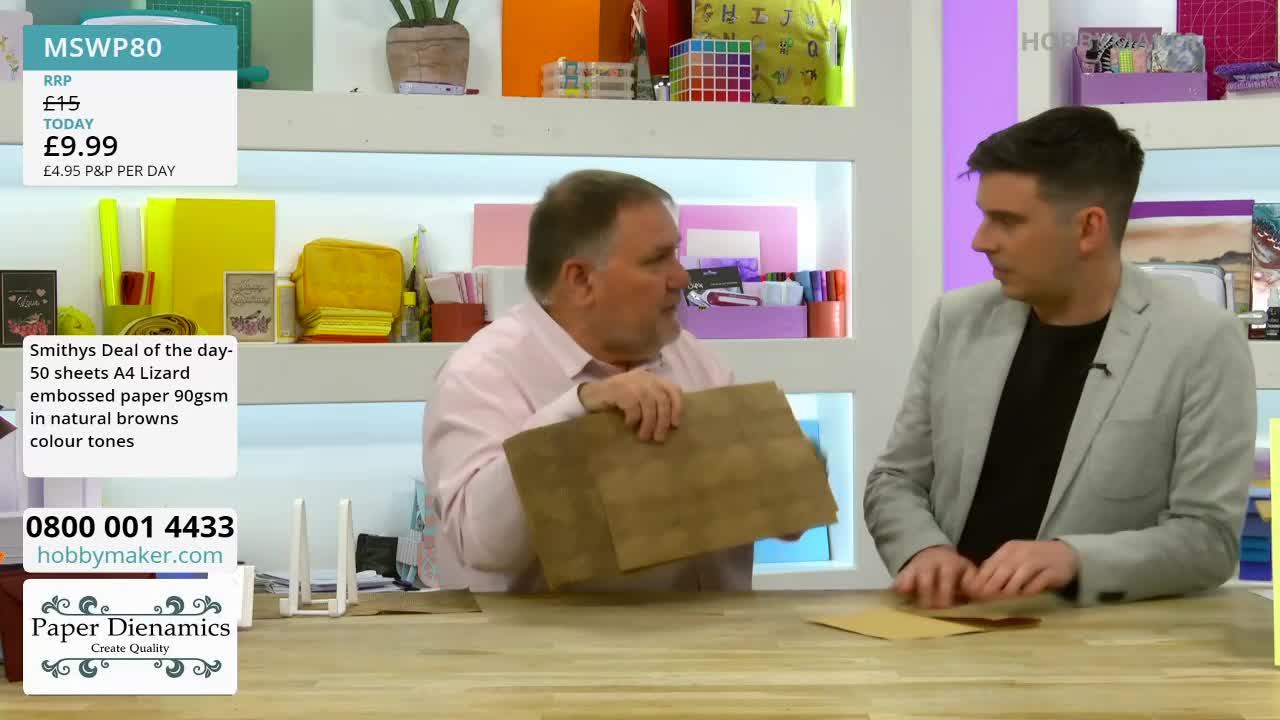 A man in a pink shirt holds up a sheet of textured brown paper. Beside him, another man in a grey jacket gestures towards a table.