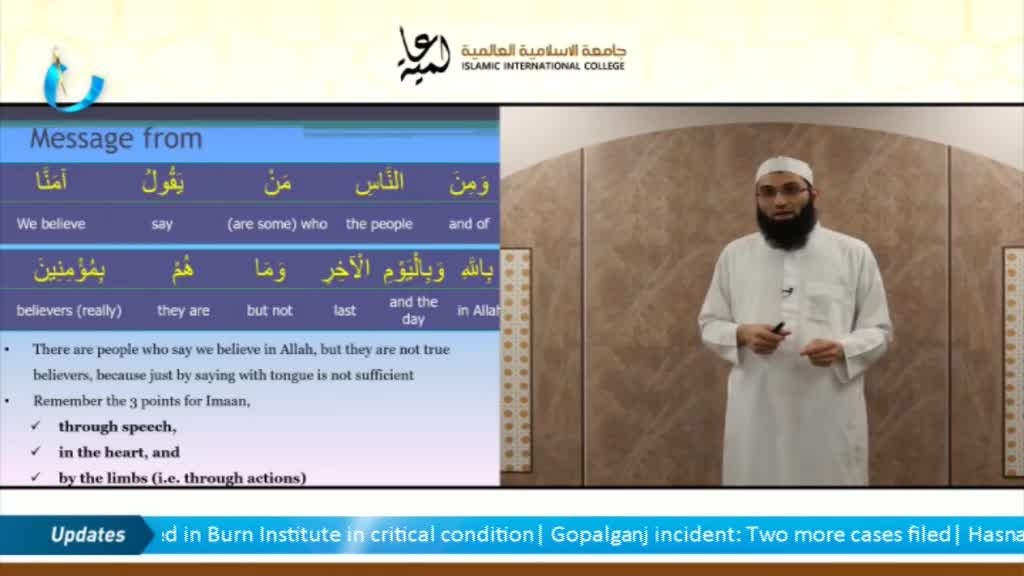 A man in a white robe stands before a wall, gesturing as he speaks. On a screen beside him, a blue and white graphic displays Arabic text and English translations.
